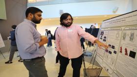 Photo of male student in a pink shirt in the center of the photo pointing to a poster of his research as another male student in a blue shirt on the left side of the photo looks on.