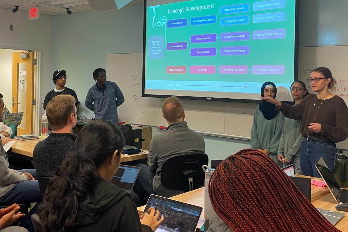 A classroom with a group of students looking at a projection screen. A group of male and female students stand at the front of the class next to the screen presenting.