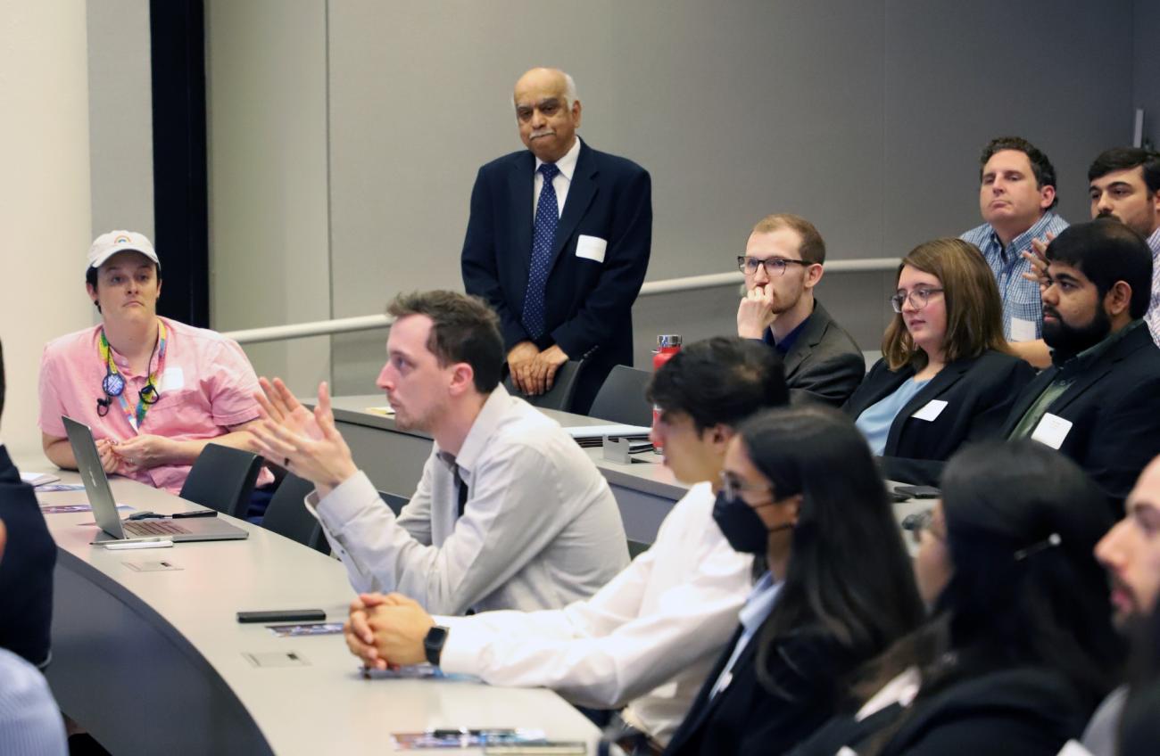 Photo of audience of people watching project teams present. Man in center sitting asking questions, while professor stands in the back listening.