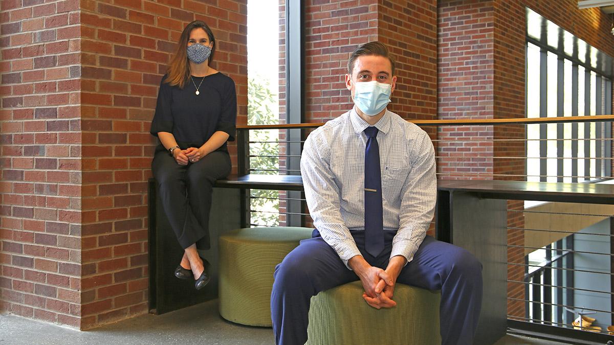 Photo of male student Joshua Lewis in the foreground sitting on a green cushioned stool while Prof. Melissa Kemp, female, sits on a small tabletop behind him.