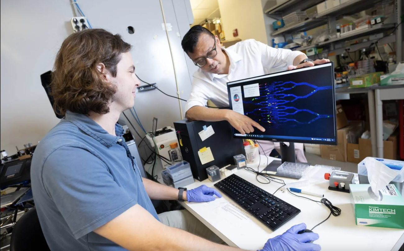 A male student with a blue shirt sits in front of a computer moving the computer mouse as a male faculty member, standing behind the computer screen, points to a digital DNA sequence on the screen.